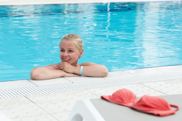 A blonde young woman in a swimming pool with red bikini left by the pool