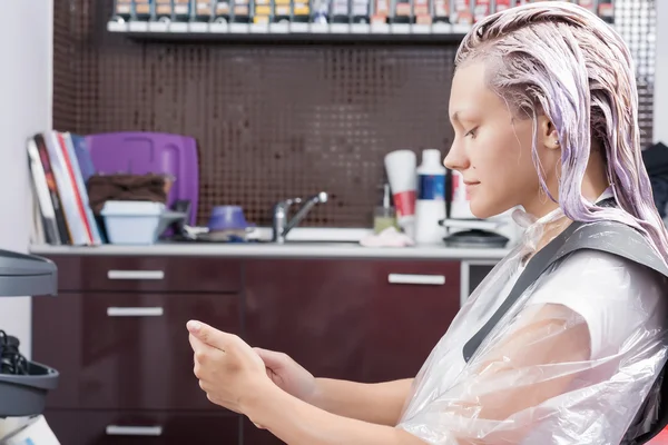 A blonde woman surfing the net with her smartphone during hair colouring
