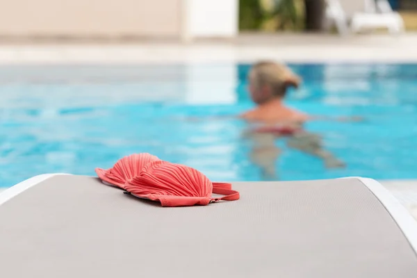 Red bikini left by a swimming pool with woman swimming in it. Selective focus