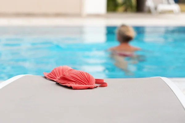 Red bikini left by a swimming pool with woman swimming in it. Selective focus