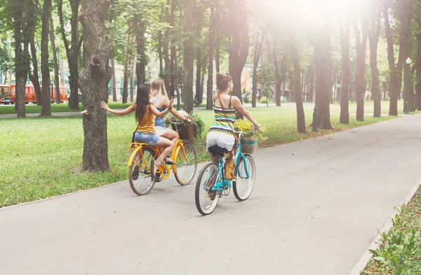 Happy boho chic girls ride together on bicycles in park