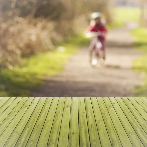 Empty, green, wooden table with small child in safety helmet on bicycle in blurred background. Ready for product display montage.