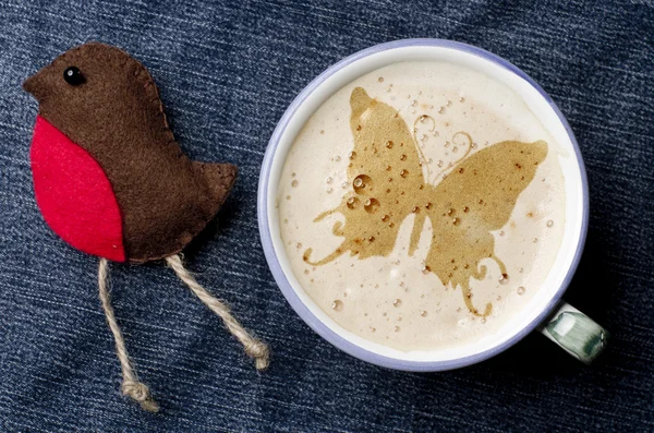 Cup of cappuccino coffee with foam in the form of butterfly on blue jeans, denim background. Robin craft handmade from felt.