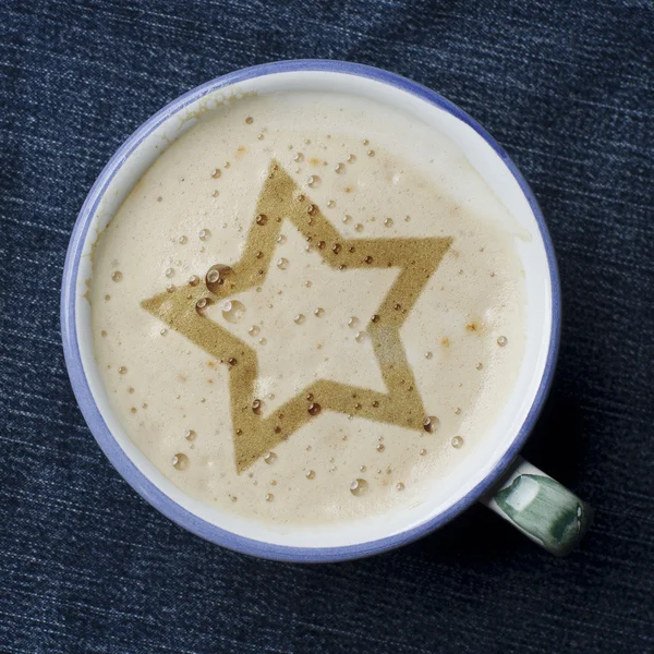 Cup of cappuccino coffee with foam in the form of star on blue jeans, denim background. White wooden star.