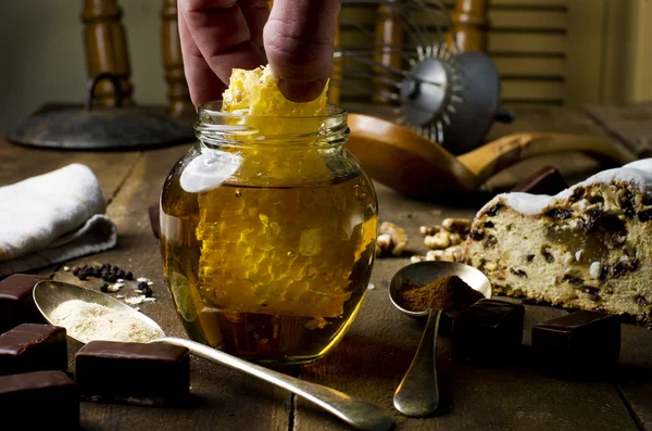 Hand reaching honeycomb from jar of healthy real honey and fruit cake on real wooden table.