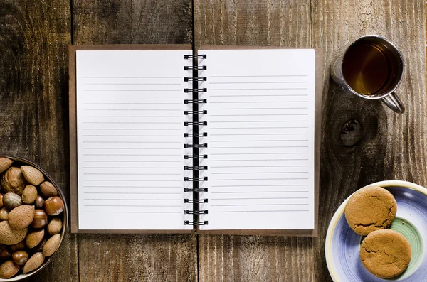 An open notebook with blank pages on wood table.Cup of tea, nuts and biscuits snack.
