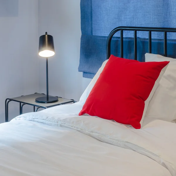 Red pillow on white bed with blue curtain and black lamp on tabl