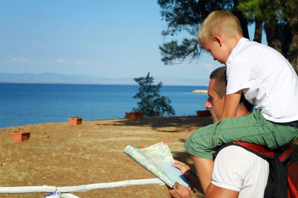 Father giving his son piggyback ride outdoors. Man and boy son looking at map infront of sea, active summer holiday vacation, family travel photo