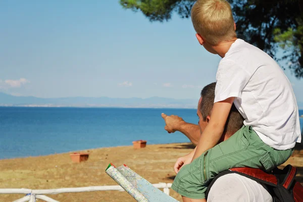 Father giving his son piggyback ride outdoors. Man and boy son looking at map infront of sea, pointing away, active summer holiday vacation, family travel photo