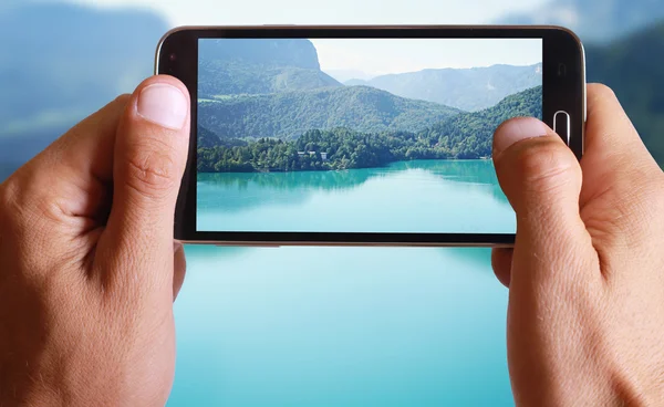 Male hand taking photo of a lake with green mountains landscape with cell, mobile phone.