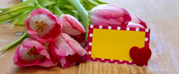 Pink tulips with heart card on wood background.