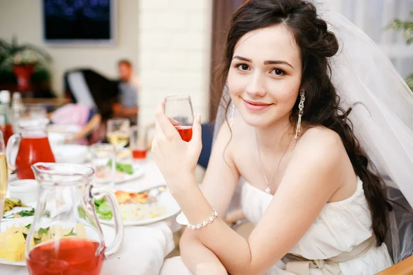 Attractive beautiful young bride in white dress sits at table with glass of red wine posing, kind eyes, smiling slightly. Celebration, wedding reception restaurant, food is on the table.