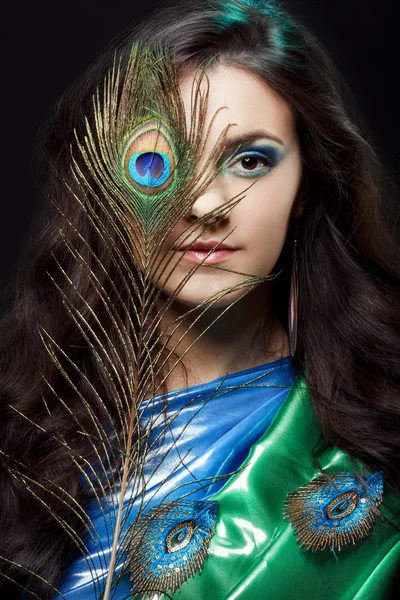 Beauty portrait of beautiful girl covering the eyes with peacock feather. Creative makeup peafowl feathers. Attractive mysterious lady. Bright harmonious colors, sari dress, shiny satin silk fabric.