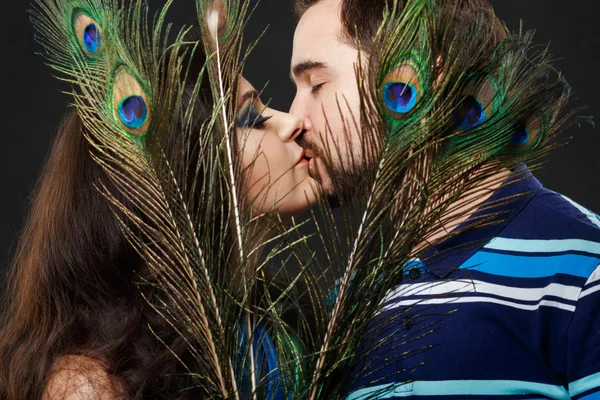 Mysterious stranger girl with long eyelashes passionately kisses a regular guy hiding behind peacock feathers. Attractive young woman in luxurious sheen, shiny fabric. Imagination, dream, fantasize.