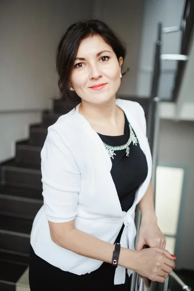 Pretty brunette business woman with kind eyes leaning forward posing on stairs of office building, wearing white jacket over tight black dress. Positive look, candid portrait. Consultations, support.