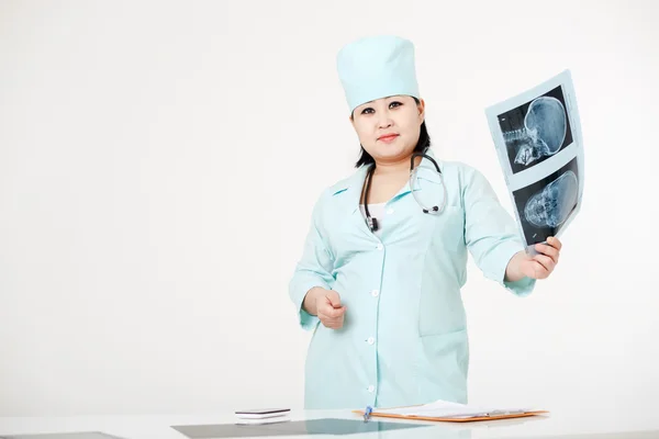 Curvy female doctor standing at table with X-ray examination results, image of skull in hand, wearing medical gown and cap, stethoscope. Asian, dark short hair. Radiology department.