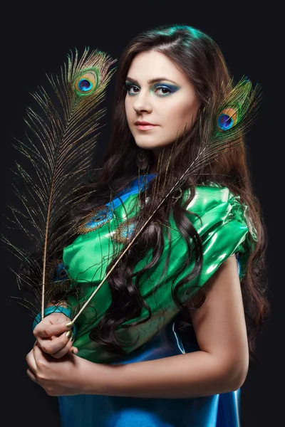 Close up beauty portrait of beautiful girl with peacock feather. Creative makeup peafowl feathers. Attractive mysterious lady. Bright harmonious colors, sari dress, shiny satin silk fabric.