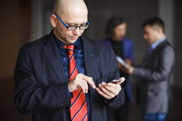 Portrait of male with glasses bald businessman using phone in hand, dressed striped suit and red tie, stands indoors office building. At background man discussing.