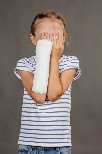 Sad girl with broken arm is standing on the gray background.