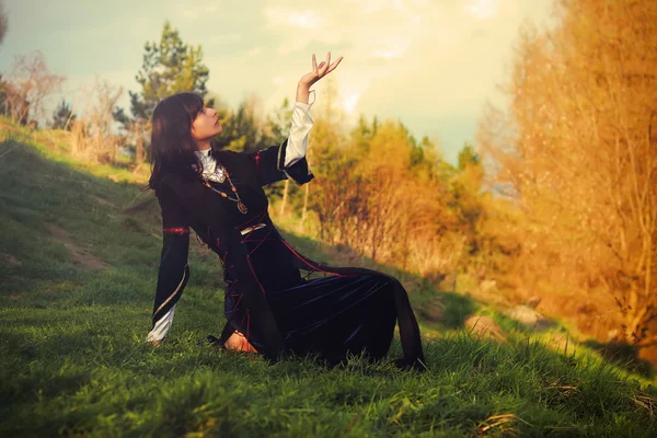 A beautiful young woman with dark hair and a historical dress posing on a meadow in open lanscape with a gesture of connection between heavenly and earth worlds
