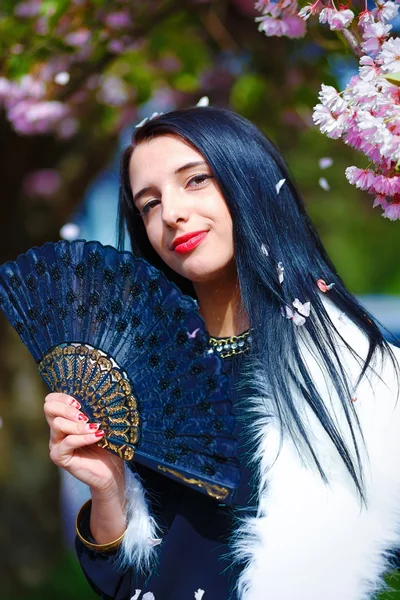 Beautiful Girl with flowers, glamour white fur and black fan in hand, posing next to blooming magical spring rosa sakura flowers. Flower background