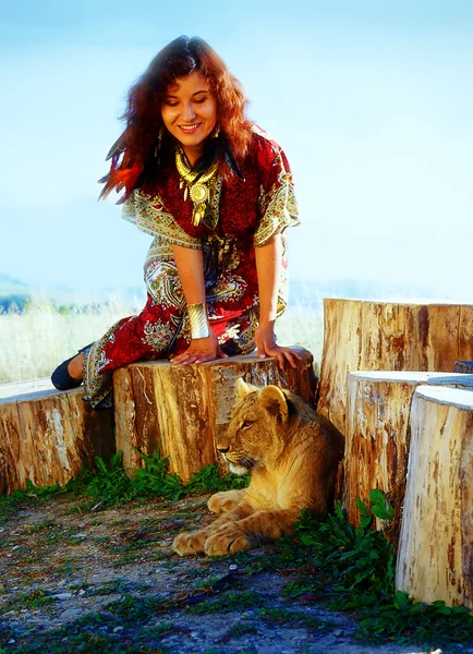 Young woman with ornamental dress and gold jewel playing with lion cub in nature.