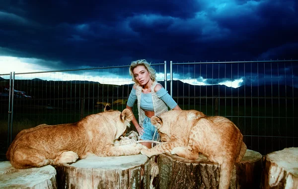 Sexy blonde woman playing with lion cub on background with beautiful blue sky and storm clouds.