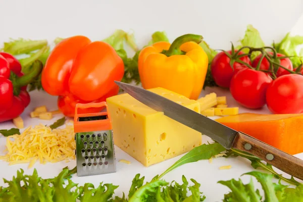 Pieces of cheeses, grated cheese, metal grate for preparing  grated cheese, knife,  tomatoes, peppers and leaves of frillis and arugula