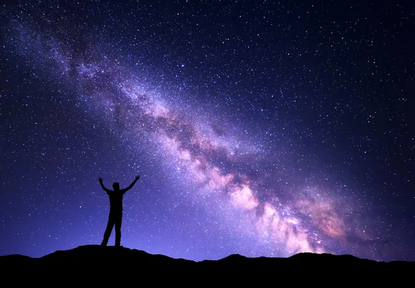 Night colorful landscape with purple Milky Way and silhouette of a standing sporty man with raised up arms on the mountain. Sky full of stars, space..
