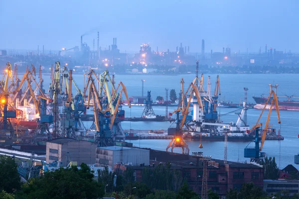 Sea commercial port at night against working steel factory in Mariupol, Ukraine. Industrial view. Cargo freight ship with working cranes bridge in sea port at twilight. Cargo port, logistic