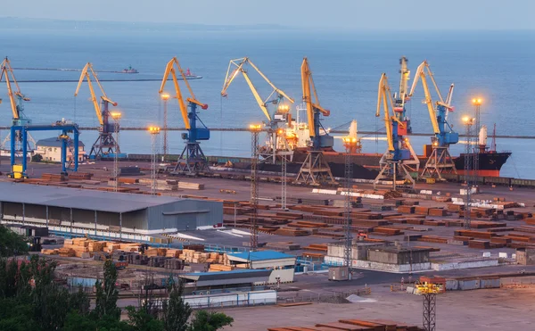 Sea commercial port at night against working steel factory in Mariupol, Ukraine. Industrial view. Cargo freight ship with working cranes bridge in sea port at twilight. Cargo port, logistic