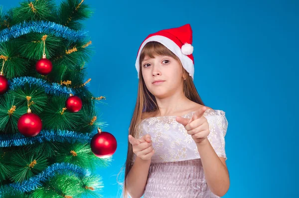 The girl with a red cap on the head costs near a beautiful New Year tree and shows forefingers forward.