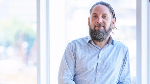 Senior man with beard looking at camera standing by window