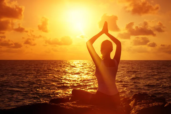 Young healthy woman is practising yoga in lotus pose at a stone at summer sunny sunrise background.