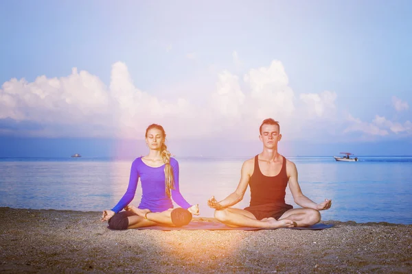 Young couple is practicing yoga meditation at summer sunrise at sea background.