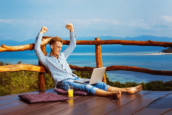 Young successful man is sitting with laptop at mountain restaurant background and raising hands in happy mood.