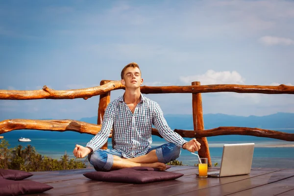 Happy young man is sitting in yoga pose with closed eyes at wooden restaraunt at natural summer mountain view background.