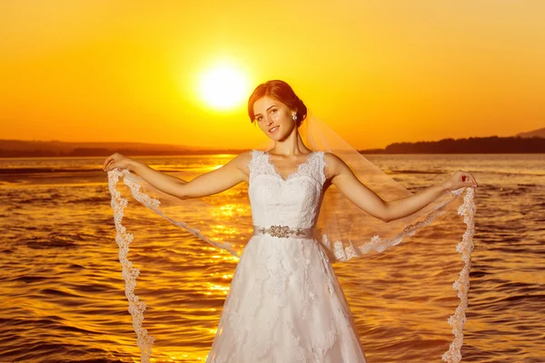 Beautiful bride in long white wedding dress is holding veil at amazing golden sea sunrise background.