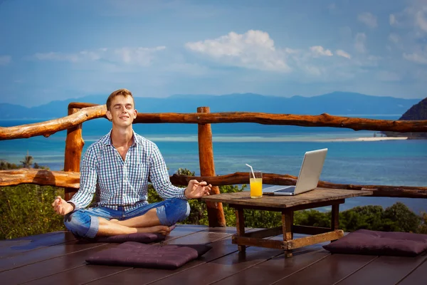 Young man with laptop at table is sitting with closed eyes in yoga pose and relaxing at a mountain restaurant at summer tropical background.