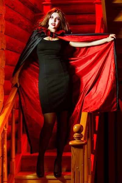 Vampire woman in cloak is standing indoors at stairs with red light background. Halloween holiday concept.