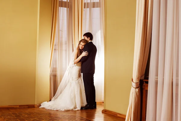 Beautiful elegant wedding couple is embracing tenderly in arch at curtained window and yellow walls background in a stunning vintage hall. Love and happy family concept.