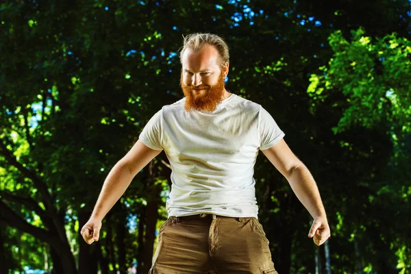 Macho man with red beard is smiling and putting hands down showing atrraction at green summer outdoor background.