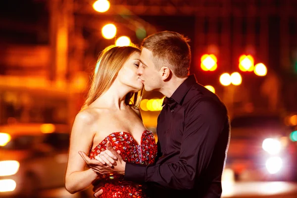 Beautiful young woman in red sparkling dress is passionately kissing elegant macho man at city street at bright lights blurred background.