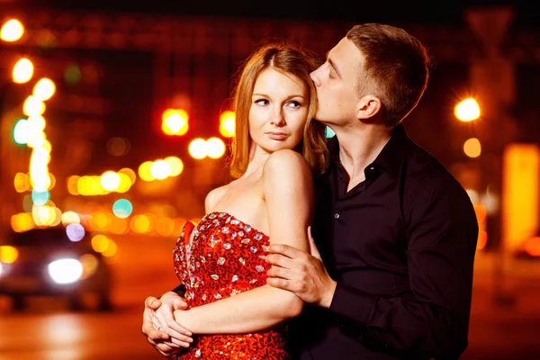 Handsome young man is embracing beautiful happy woman in red dress at yellow street lights background.
