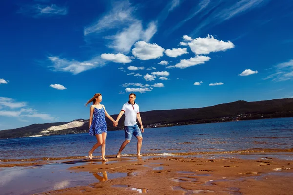 Beautiful young couple is walking along  river coast with yellow sand at blue summer cloudy sky background.