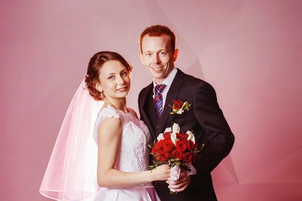 Portrait of happy smiling wedding couple holding red roses flowers bouquet at pink wall background.