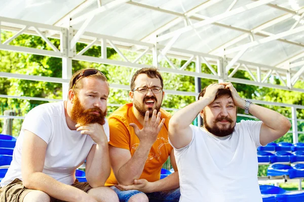 Three bearded football fan friends are sitting upset at goal moment at summer stadium  background.