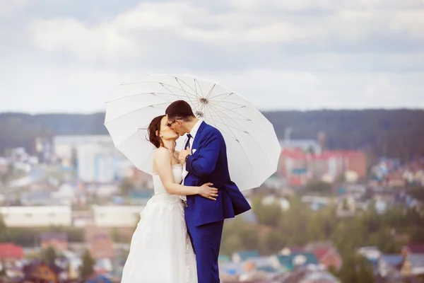 Young happy wedding couple with white umbrella is kissing at hill top at blue sky view background.