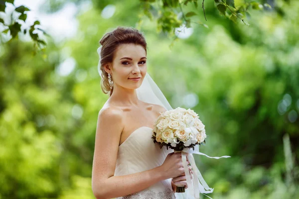 Closeup portrait of beautiful bride with flowers bouquet standing outdoors at green natural background.