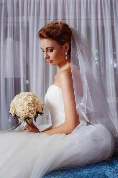 Vertical portrait of beautiful young bride holding roses wedding bouquet at white curtain background.